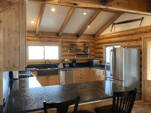 A rustic kitchen with log walls, granite countertops, stainless steel appliances, and wooden beams.