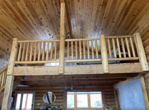 Interior of a rustic cabin featuring wooden beams, a loft with a railing, and large windows letting in natural light.