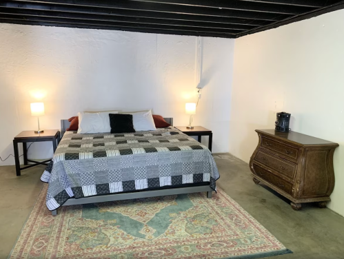 A simple bedroom in a basement with a bed, two lamps, a dresser, and a coffee maker on a side table.