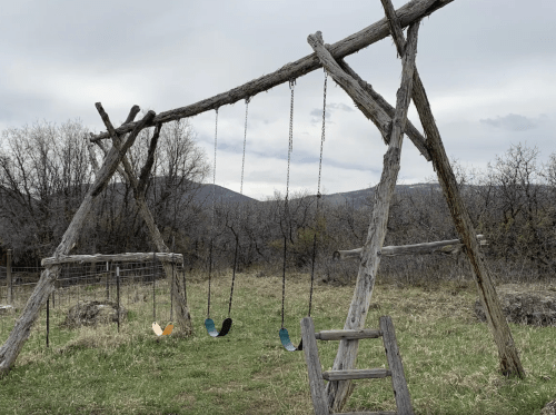A rustic wooden swing set with four swings, set against a backdrop of trees and mountains under a cloudy sky.