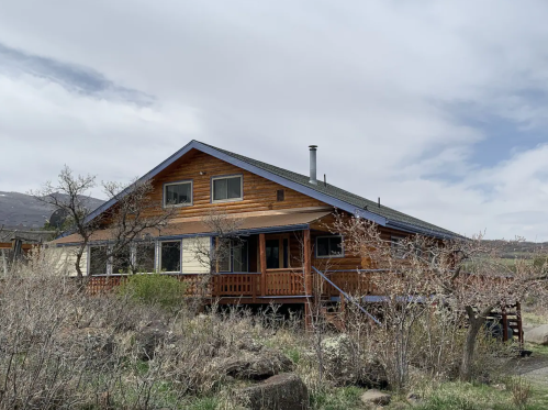 A rustic wooden house with a porch, surrounded by sparse vegetation and a cloudy sky in the background.