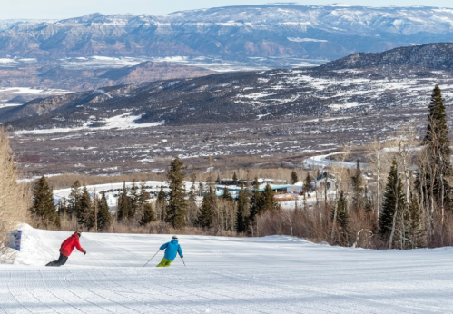Two skiers descend a snowy slope with mountains and a valley in the background on a clear day.