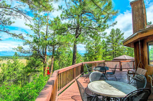 A wooden deck surrounded by pine trees, featuring outdoor seating and a scenic view of mountains and blue skies.