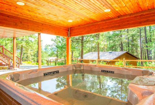 A hot tub under a wooden porch, surrounded by trees and a cabin in the background. Bright and inviting atmosphere.