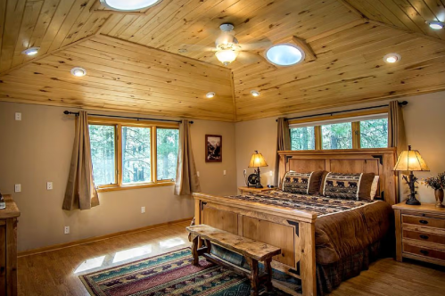 Cozy bedroom with wooden ceiling, large windows, a bed with patterned bedding, and warm lighting.