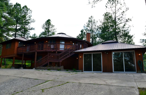 A modern wooden house with a circular design, surrounded by tall trees and featuring a large deck and glass doors.