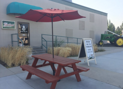 A red picnic table with an umbrella outside a building, with a sign and a green tractor nearby.