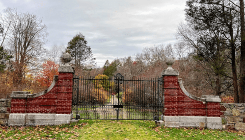 A wrought iron gate with brick pillars opens to a tree-lined path, surrounded by autumn foliage.