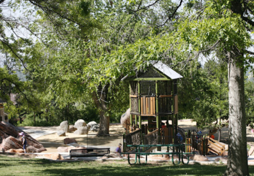 A playground with a wooden structure surrounded by trees and rocks, with children playing in the background.