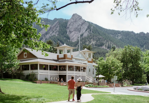 A couple walks on a path near a historic building surrounded by trees and mountains on a sunny day.