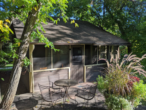 A small, rustic cabin surrounded by greenery, featuring a patio with a table and chairs.