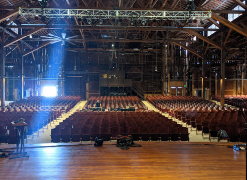 A spacious auditorium with rows of empty seats, wooden beams, and a stage, illuminated by a beam of light.