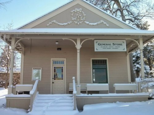A quaint general store with a sign, surrounded by snow, featuring a porch and decorative architectural details.