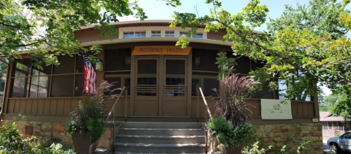 Front view of a charming building labeled "Missions House," surrounded by greenery and featuring an American flag.