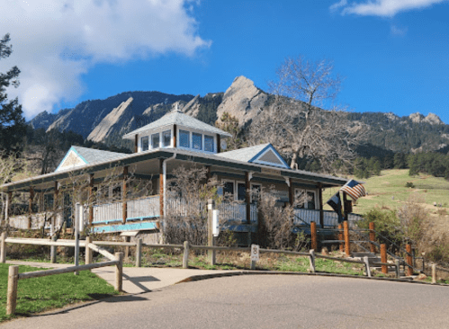 A charming building with a porch, surrounded by trees, set against a backdrop of rocky mountains and blue sky.