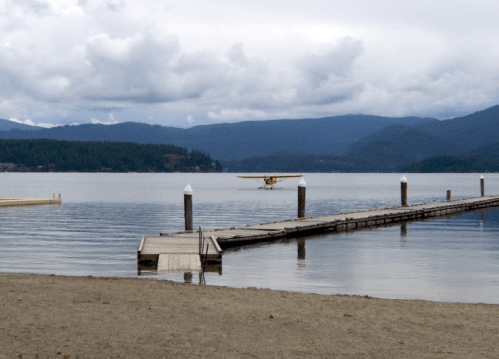 A seaplane lands on a calm lake near a wooden dock, surrounded by mountains and cloudy skies.