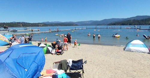 A sunny beach scene with families enjoying the water, umbrellas, and a pier in the background. Mountains are visible.