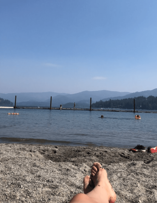 A sandy beach with a person relaxing by the water, a dock in the distance, and mountains under a clear blue sky.