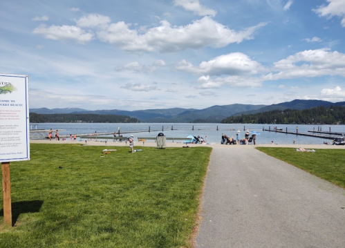 A scenic view of a lake with people enjoying the beach, surrounded by mountains and a blue sky with clouds.