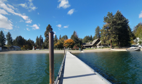 A wooden dock extends into a calm lake, with trees and a beach visible in the background under a clear blue sky.