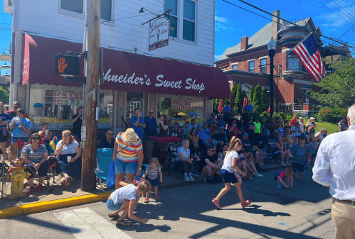 A crowd gathers outside Schneider's Sweet Shop, enjoying a sunny day during a local event or parade.