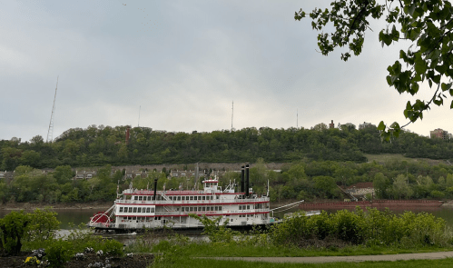 A riverboat with a red and white design sails past a green hillside under a cloudy sky.