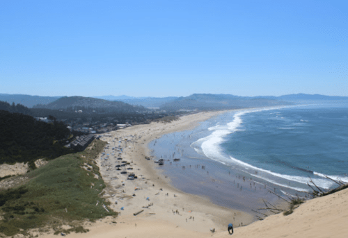 A scenic view of a sandy beach with gentle waves, surrounded by hills and a clear blue sky.