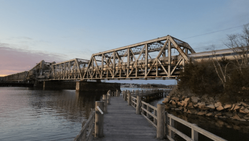 A metal bridge spans a calm waterway at sunset, with wooden walkways leading towards it.