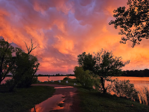 A serene lakeside path at sunset, with vibrant orange and purple clouds reflecting on the water.