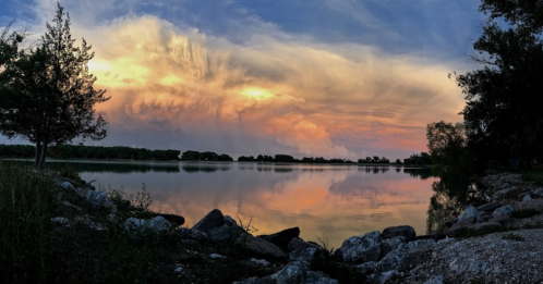 A serene lake at sunset, reflecting colorful clouds and surrounded by trees and rocky shoreline.