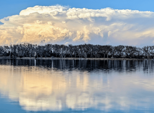 A serene landscape featuring a calm lake reflecting fluffy clouds and trees along the shore under a blue sky.
