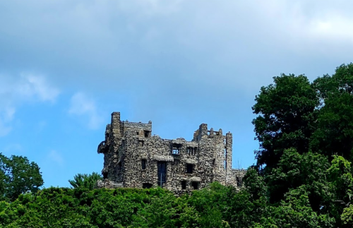 Ruins of a stone castle atop a green hill, surrounded by trees under a partly cloudy sky.