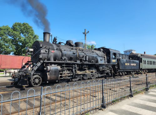 A vintage steam locomotive with smoke billowing, labeled "New Haven," parked beside a train station on a sunny day.