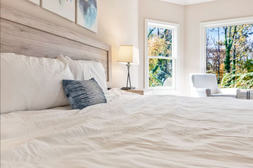 Cozy bedroom with a light-colored bed, decorative pillow, and large windows showcasing a natural view.