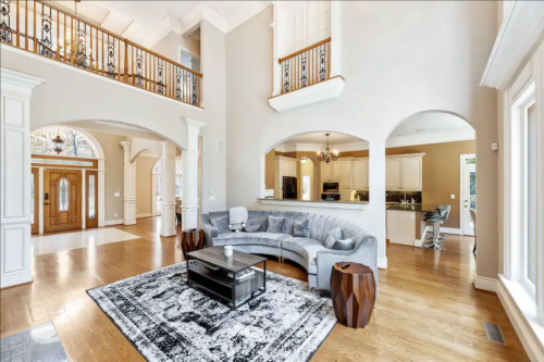 Spacious living room with a curved gray sofa, wooden floors, and large windows, leading to a kitchen area.