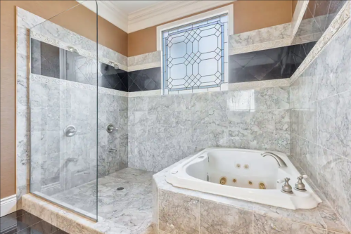 Luxurious bathroom featuring a glass shower, marble walls, and a jetted tub with a decorative window.