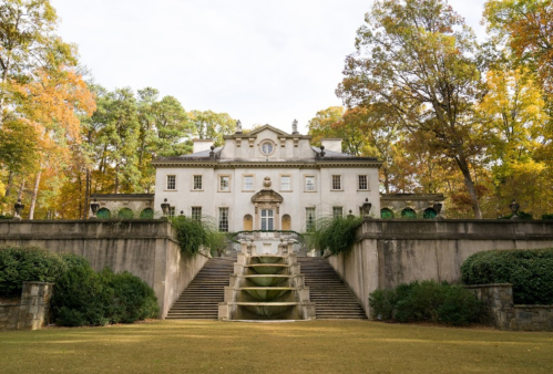 A grand, historic mansion with a staircase and lush greenery, surrounded by colorful autumn trees.