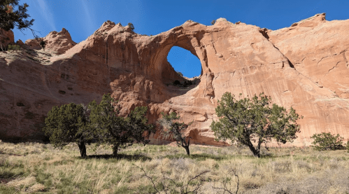 A large rock formation with a natural arch, surrounded by sparse vegetation and blue sky.