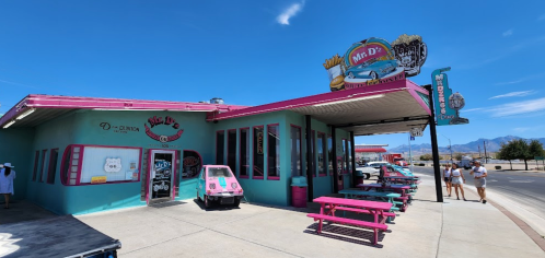 Colorful diner exterior with pink accents, outdoor seating, and a vintage car display under a clear blue sky.