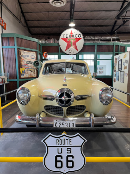 A vintage yellow car displayed in a museum, with a Route 66 sign and Texaco logo in the background.