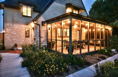 A cozy home exterior featuring a screened porch with string lights, surrounded by lush gardens at dusk.