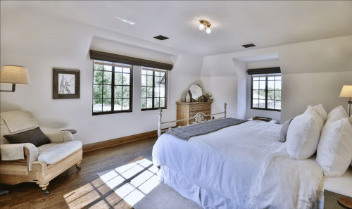 Bright, airy bedroom with a large bed, armchair, and windows, featuring wooden floors and minimalist decor.