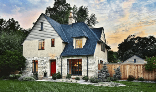 A charming two-story house with a stone facade, dark roof, and landscaped yard, set against a colorful sunset sky.