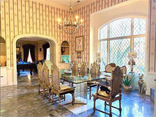 Elegant dining room with a glass table, ornate chairs, and patterned wallpaper, featuring large windows and decorative accents.