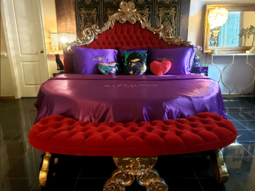 Luxurious bedroom featuring a red and gold bed with purple bedding and decorative pillows, set against a stylish backdrop.