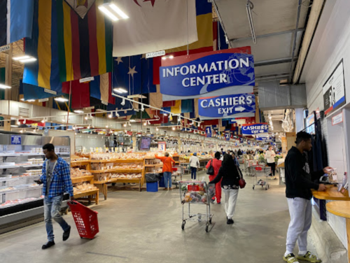 A bustling market interior with flags hanging, shoppers browsing, and signs for the information center and cashiers.