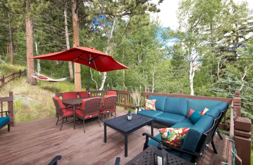 Cozy outdoor deck with seating, a table, and a red umbrella, surrounded by trees and greenery.