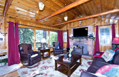 Cozy living room with wooden beams, a stone fireplace, and large windows overlooking greenery. Comfortable seating and decor.