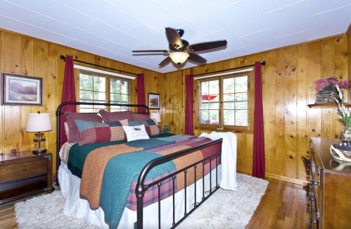 Cozy bedroom with wooden walls, a metal bed, colorful bedding, and large windows letting in natural light.
