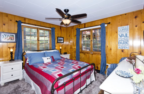 Cozy bedroom with wooden walls, a patterned quilt, two windows, and a ceiling fan, featuring a dresser and decorative items.